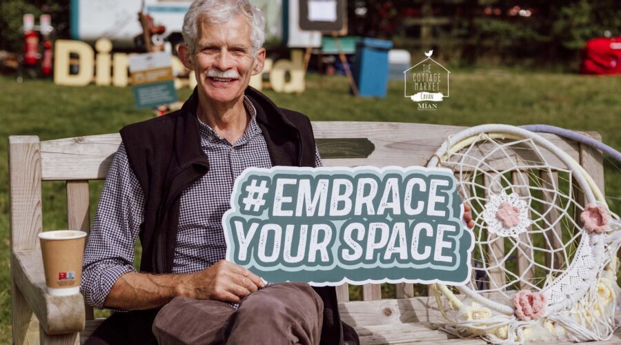 Older man holds sign that says #EmbraceYourSpace while sitting on wooden bench.