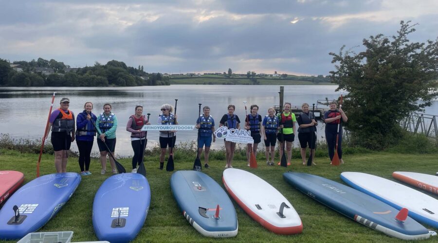 Group of women hold paddles and stand next to boards at shoreline.