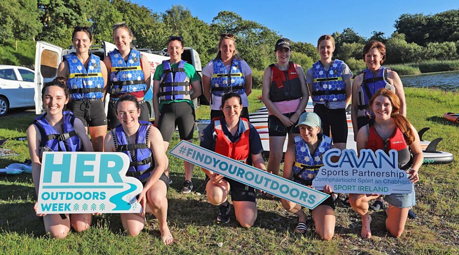 Group of women in life jackets stand near shoreline holding signs for Her Outdoors Week.